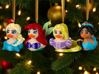 Four Disney princess ornaments hanging on a Christmas tree with lights in the background.